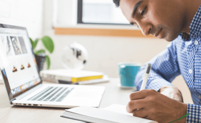 man leaning over his desk writing in a journal next to a laptop